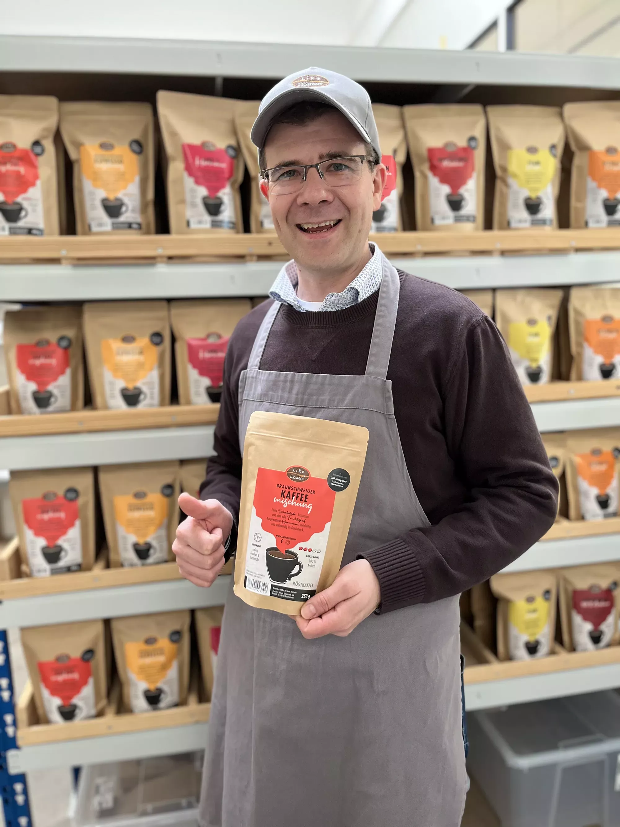 Markus Ratajczak steht vor einem Regal mit Kaffees und hält einen Kaffee in der Hand.